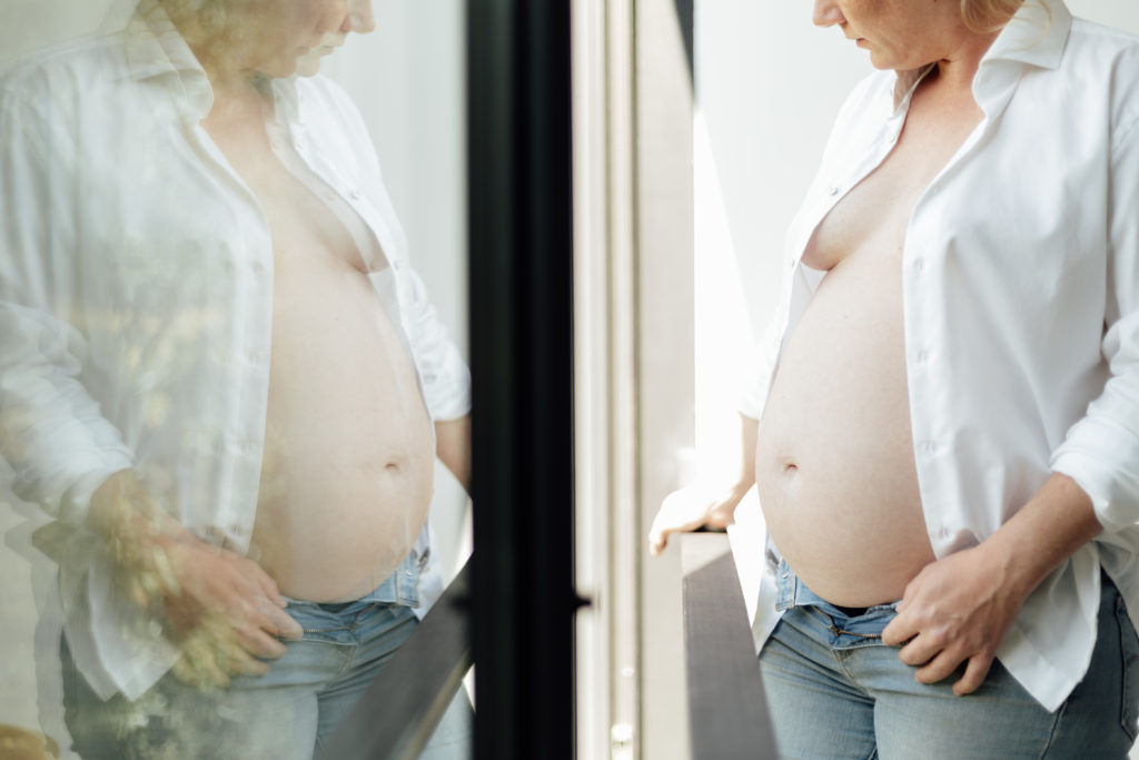 A pregnant woman stands looking at her reflection in the window
